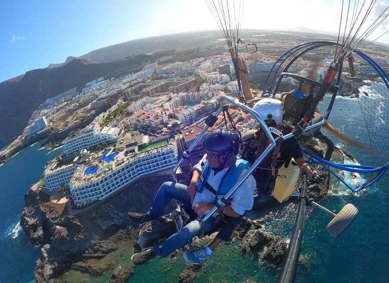 A person in a harness and helmet, seated in a cable car-like apparatus, overlooking a coastal resort area with buildings and a clear blue sea. 5
