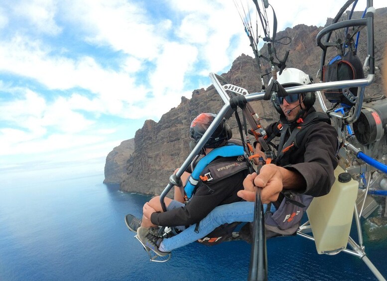 A person in a harness and helmet, seated in a cable car with a rocky landscape and ocean in the background. 1