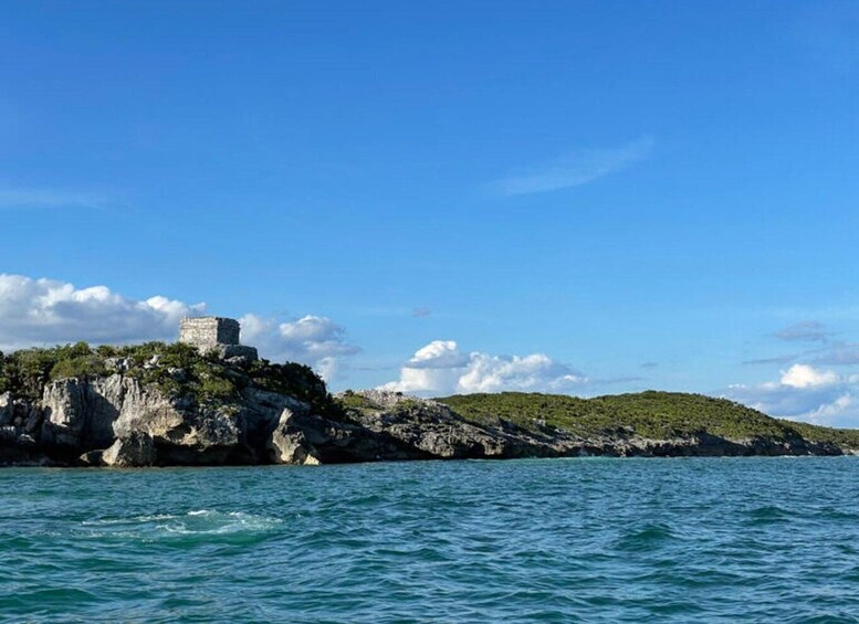A coastal landscape with a rocky outcrop and a stone structure on top, surrounded by clear blue waters and a partly cloudy sky. 1