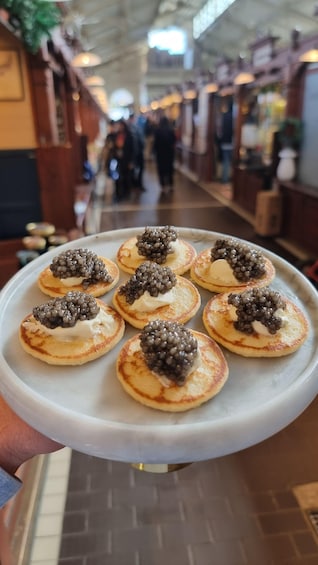 A tray of pancakes topped with caviar and cream, held by a person in a market. 1