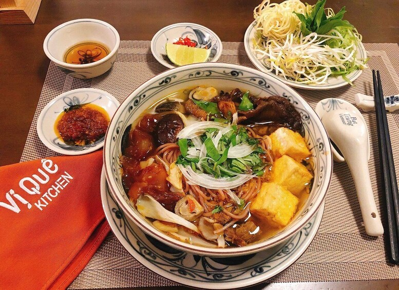 A bowl of soup with noodles, tofu, and vegetables, accompanied by a side of bean sprouts and a small dish of chili sauce. 5
