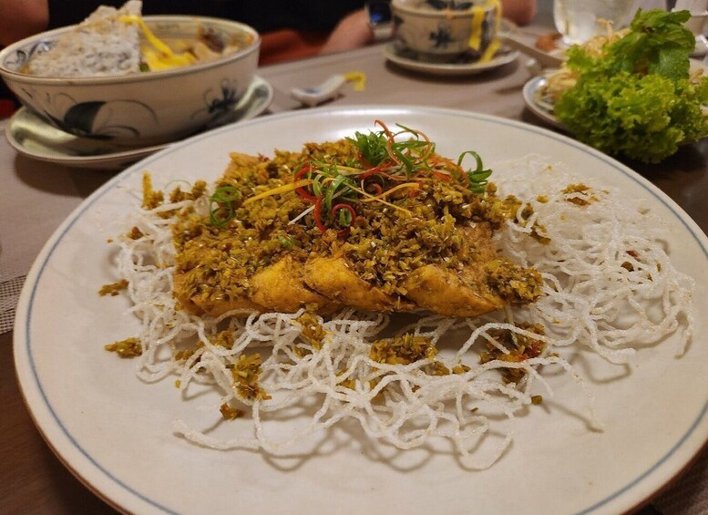 A plate of fried fish with a crispy coating, garnished with herbs and vegetables, served over a bed of white noodles. 3