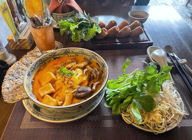 A bowl of soup with noodles and vegetables, a plate of spring rolls, a glass of iced tea, and fresh herbs on a table. 4