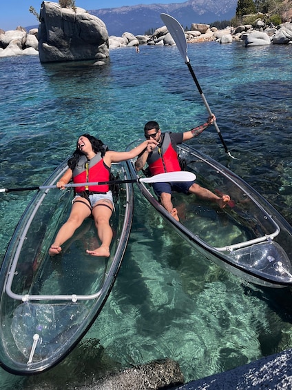 Two people in a transparent kayak on a clear lake with rocky shores and mountains in the background. 3
