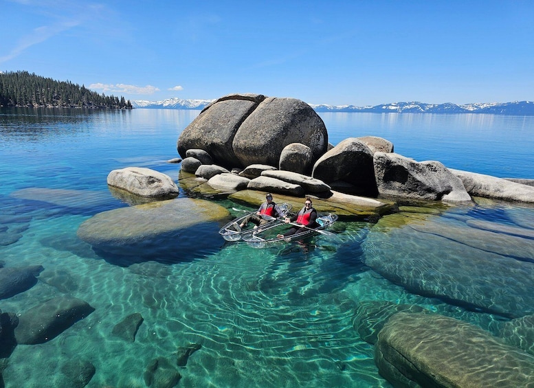 Two people in a transparent kayak on clear water with large rocks and a mountain range in the background. 1