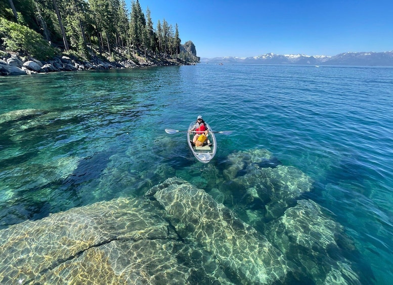 A kayaker on clear, shallow water with visible rocks and a forested shoreline. 5