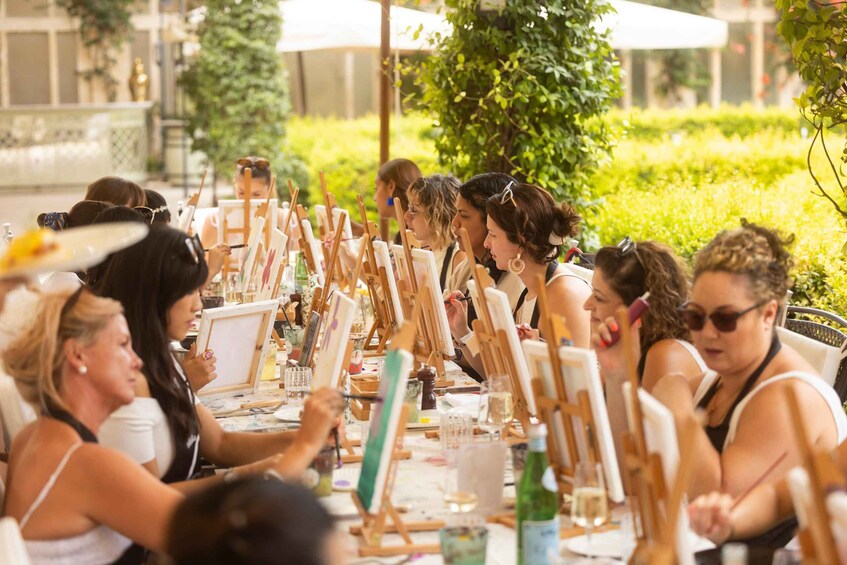 A group of people engaged in an outdoor painting activity, seated at easels with art supplies. 5