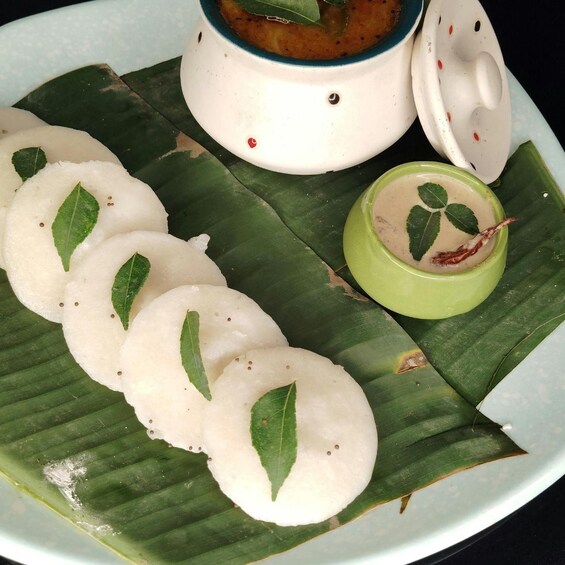 A plate of idlis with coconut leaves garnish, a bowl of sambar, and a small bowl of coconut chutney. 5