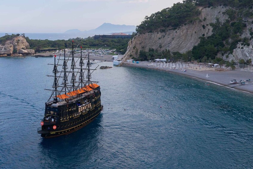 A tall ship named 'GOSTER' sailing in a bay with a beach and rocky cliffs in the background. 3