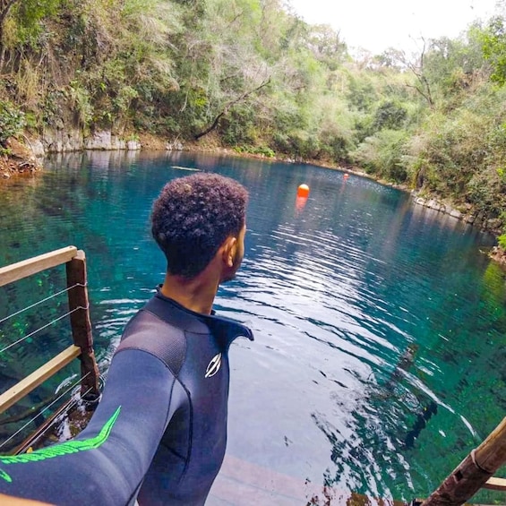 A person in a wetsuit standing on a dock by a clear blue river surrounded by greenery. 1