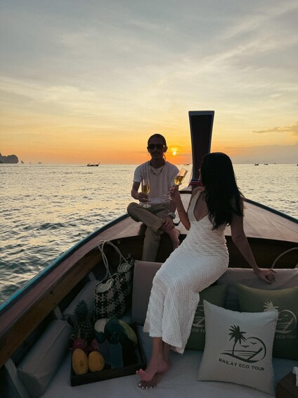 Two people on a boat at sunset with drinks, tropical fruits, and a pillow with 'RAILAY ECO TOUR' text. 2