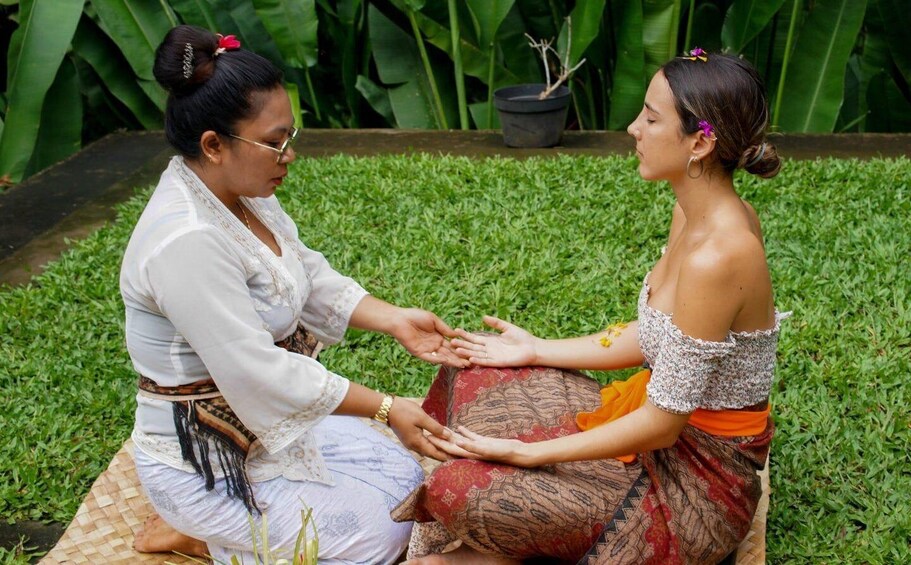 Two people sitting on a mat outdoors with greenery in the background. 1