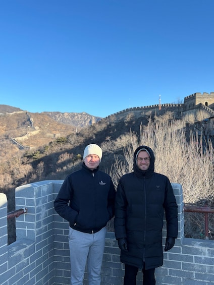 Two people standing on a brick wall with a mountainous landscape and a historical wall in the background. 2