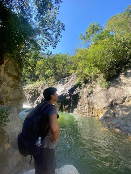 A person standing by a river with a backpack, surrounded by greenery and rocky cliffs. 2