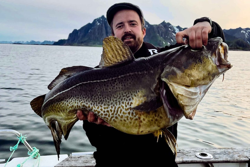 A person holding a large fish on a boat with a mountainous landscape in the background. 1