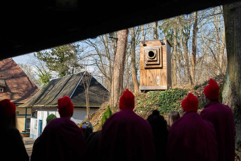 A group of people in red hats observing a wooden structure with a circular opening, possibly a birdhouse, in a wooded area with leafless trees and a house with a sloped roof. 2