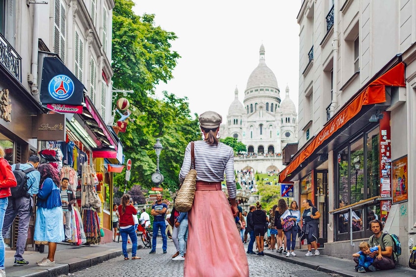 A busy street with people walking, shops, and a large dome building in the background. 1