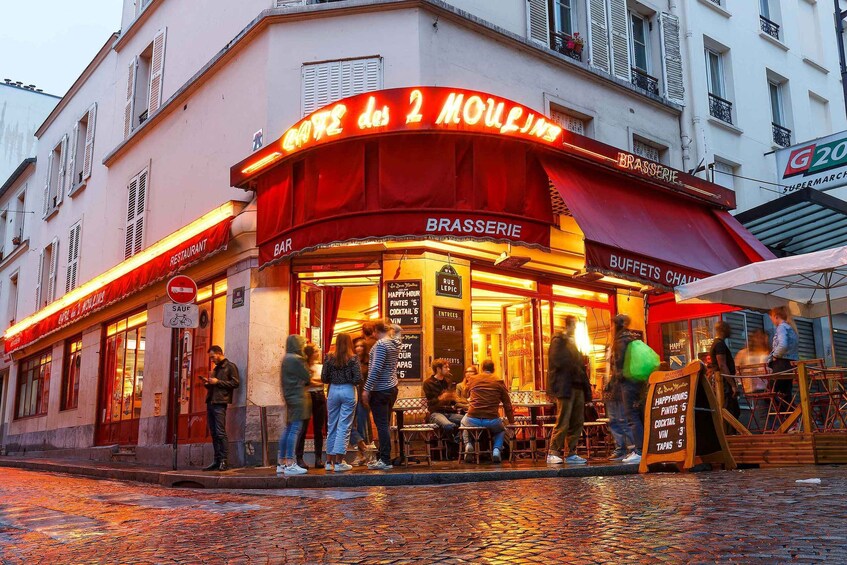 A café with a red awning and outdoor seating. People are gathered around tables, and a signboard displays menu items. 2
