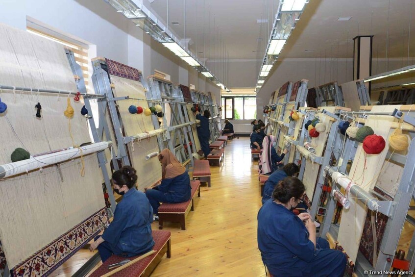 A room with people engaged in textile weaving on large looms. 1