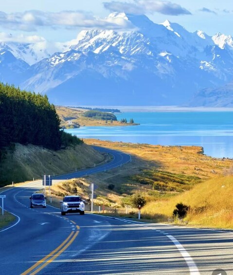A winding road with vehicles, a body of water, and snow-capped mountains in the background. 1