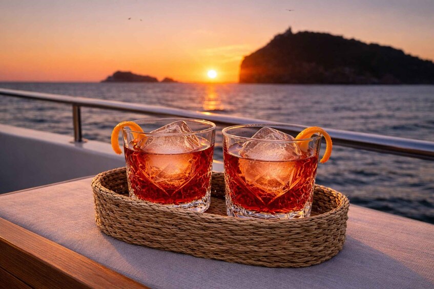 Two glasses of amber-colored drink with ice and orange peel garnish on a woven tray by a railing overlooking a sunset over the sea. 1