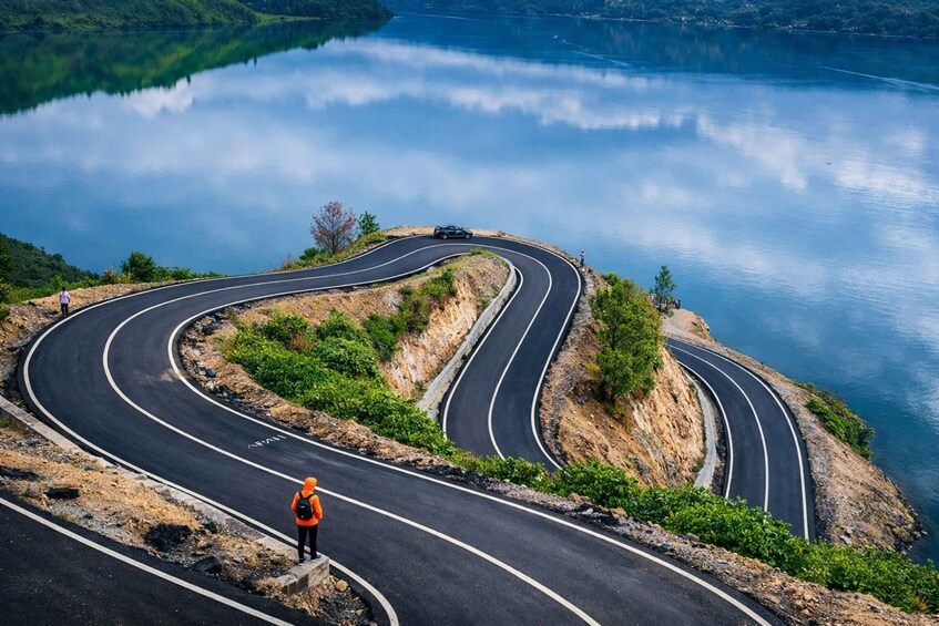 A winding mountain road with a person in orange safety gear standing on the side. 5
