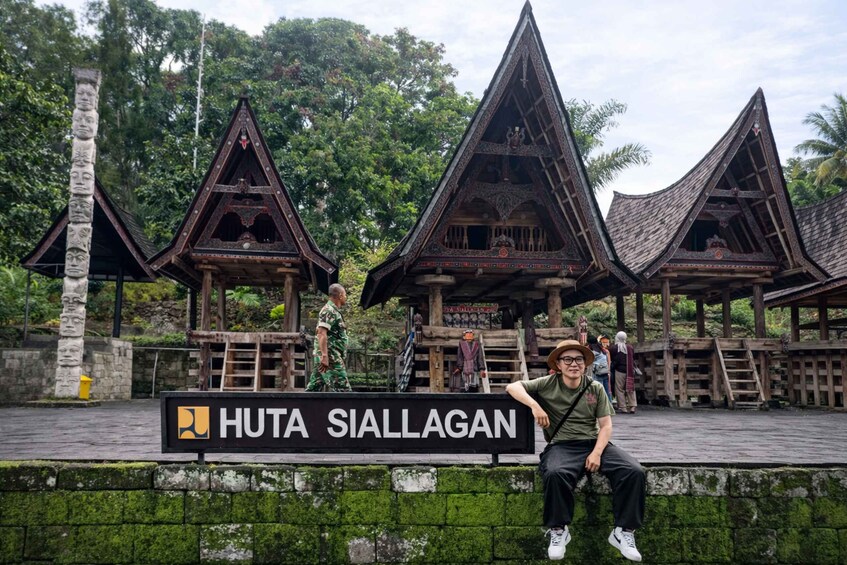A traditional wooden structure labeled 'Huta Siallagan' with a person sitting on a wall in front. 3