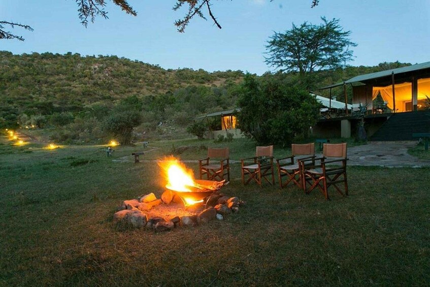 A campfire with chairs around it, a building with a porch, and a hillside in the background. 2