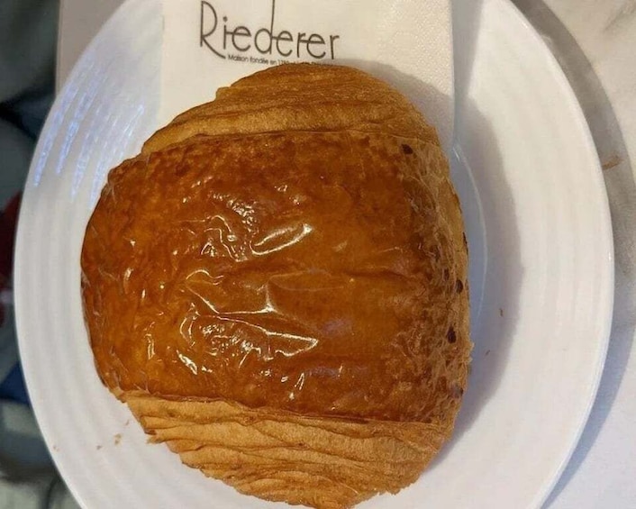 A glazed pastry on a white plate with a paper label that reads 'Riederer.' 2