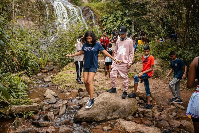 A group of people crossing a rocky stream with a waterfall in the background. 2
