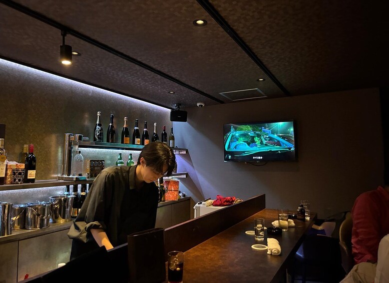 A bar with a bartender preparing drinks, bottles of wine on shelves, and a TV displaying a car scene. 1