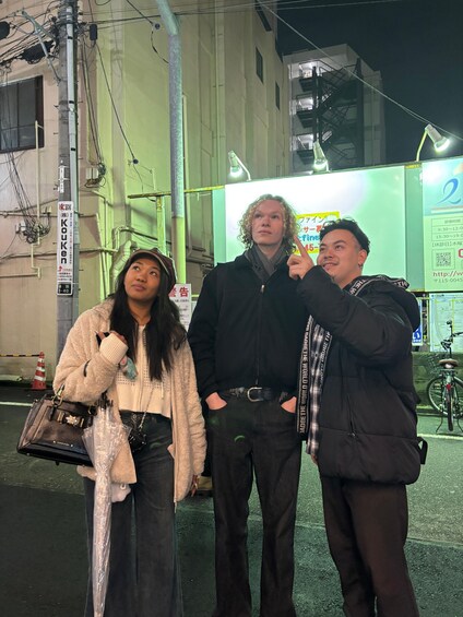 Three people standing on a street at night with buildings and a signboard in the background. 5