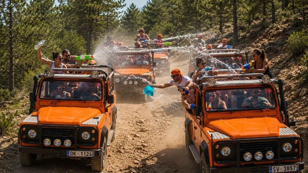 A convoy of orange off-road vehicles driving through a dirt path with people spraying water from hoses. 1