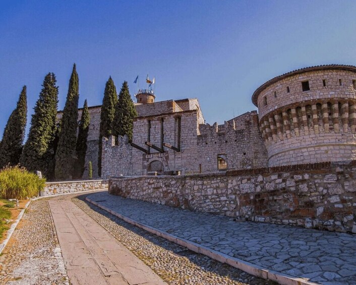 A historic stone fortress with a central tower and a round tower, surrounded by a cobblestone path and tall cypress trees. 3