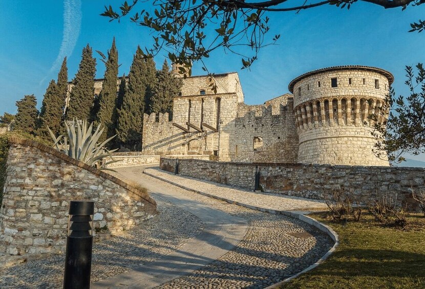 A historic stone fortress with a round tower, surrounded by trees and a gravel pathway. 2