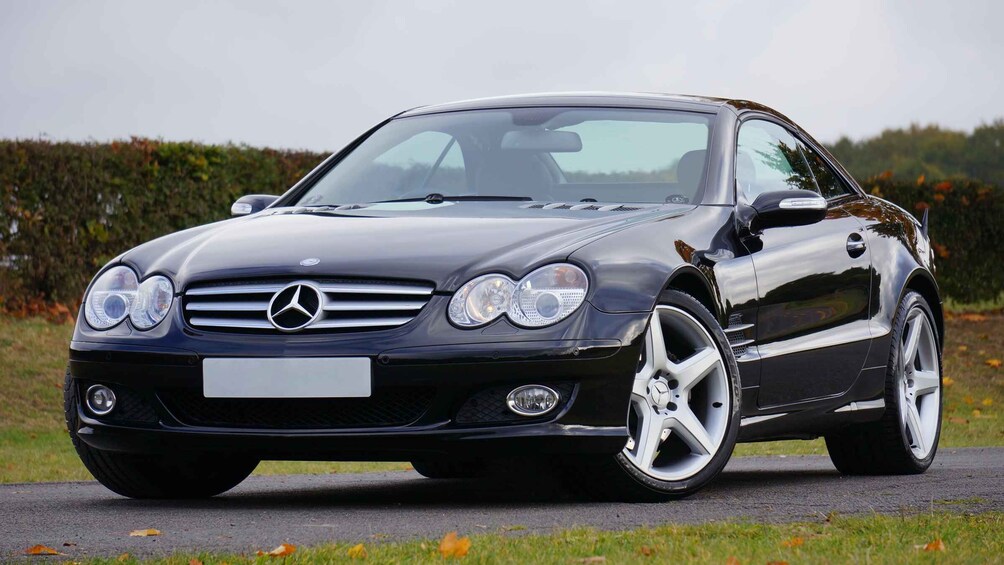 A black Mercedes-Benz with a visible license plate, parked on a road with a hedge in the background. 4