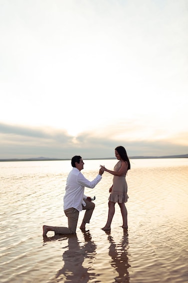 A man kneeling in shallow water, proposing to a woman standing nearby. 3