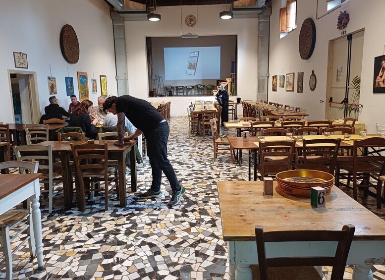 A café with mosaic flooring, wooden tables, and chairs, and a person bending over a table. 2