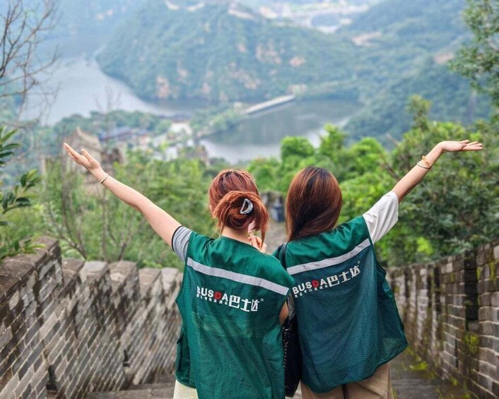 Two people wearing green shirts with 'BUSPA' text, standing with arms outstretched on a stone wall overlooking a river and mountains. 1