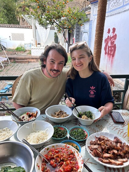 Two people sitting at an outdoor table with various dishes of food, including noodles, vegetables, and meat. 3