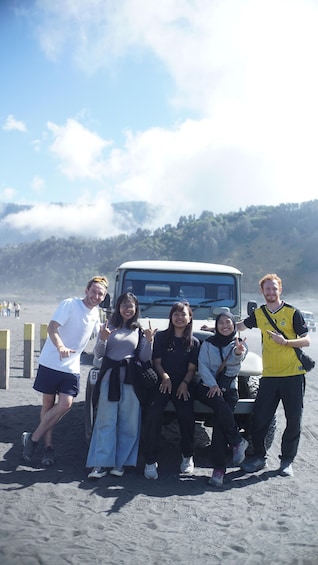 A group of people posing in front of a vehicle on a sandy beach with a forested hill in the background. 2