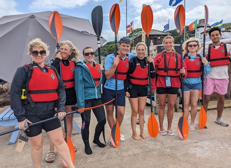 A group of people in red life jackets holding paddles, standing outdoors with flags and a tent in the background. 1