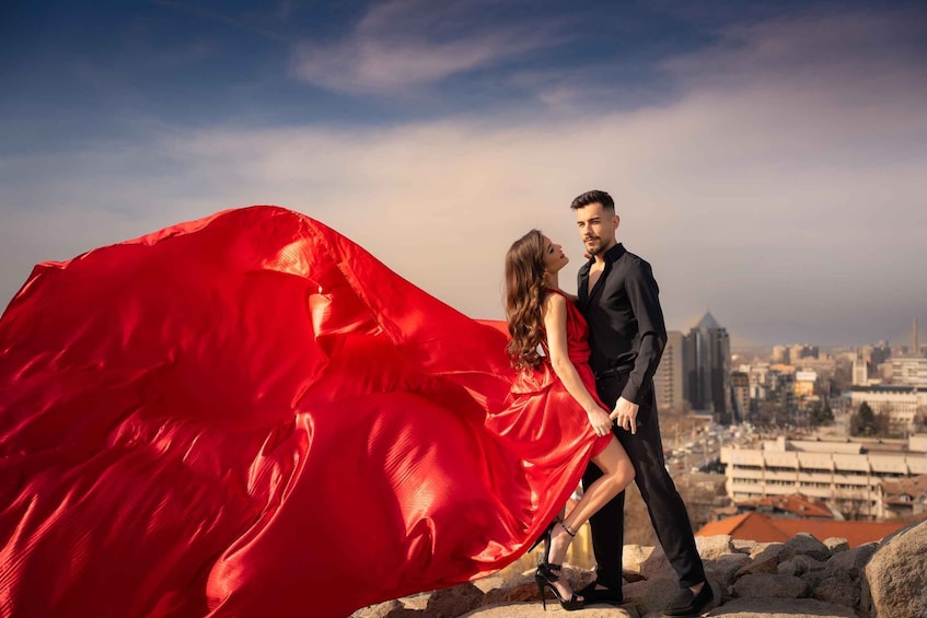 A couple standing on a rocky outcrop with a cityscape in the background. 2