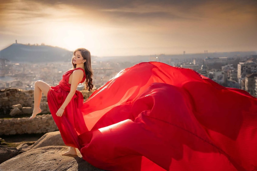 A person in a red dress sitting on a rock with a cityscape in the background. 1