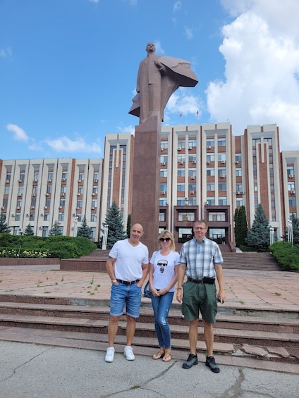 Three people standing in front of a large statue and a building with multiple windows. 2