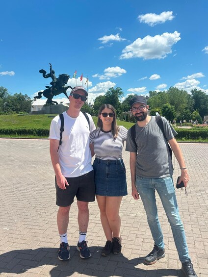 Three people standing in front of a statue with flags, in a park with trees and a clear sky. 4