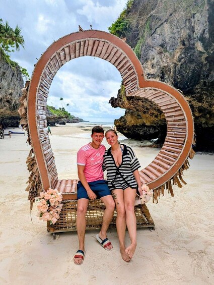 A couple sitting on a heart-shaped bench made of natural materials on a sandy beach with a rocky cliff in the background. 2
