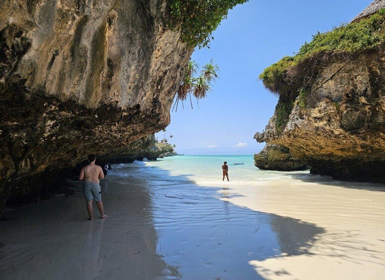 A beach with clear turquoise water, a person standing in the water, and a large rock formation on the left. 4