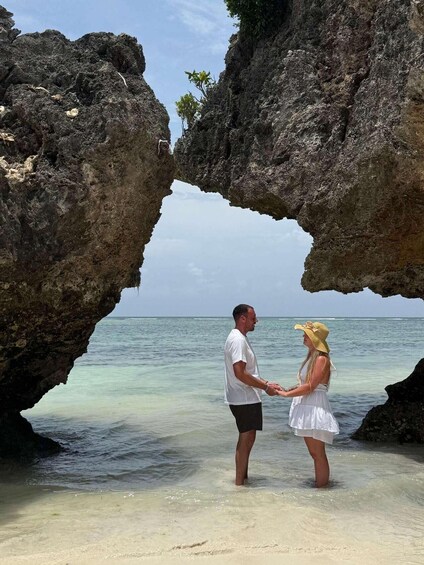A couple holding hands in a natural rock arch formation with clear turquoise water in the background. 4