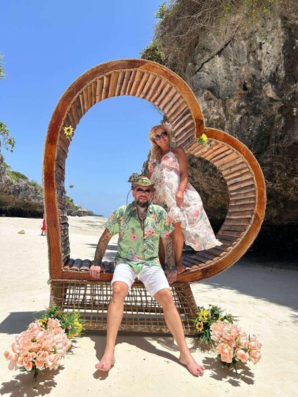 A man and a woman sitting on a wicker swing chair on a sandy beach with flowers and a rocky cliff in the background. 3